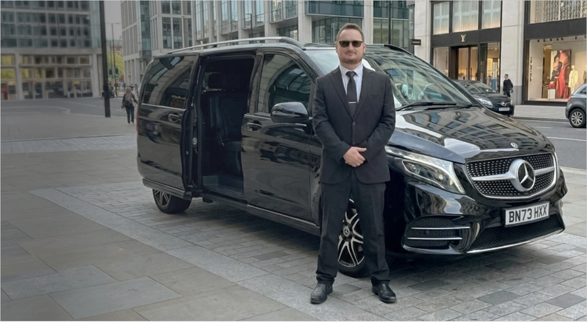 Premium chauffeur-driven Mercedes V-Class for private hire in the UK, suited driver standing beside luxury black MPV in city street